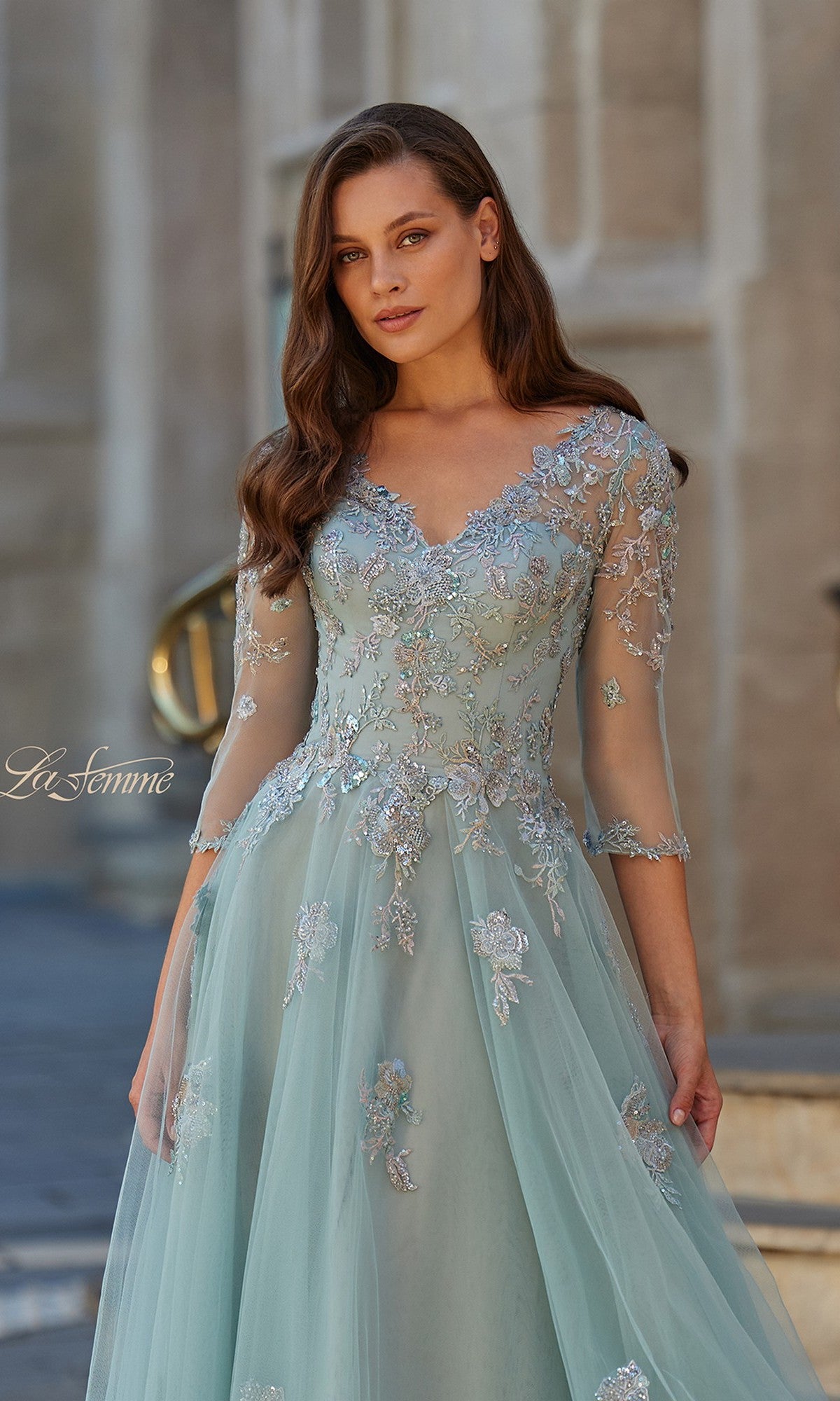 A woman wearing the La Femme 33536 long formal dress with light blue tulle, floral embroidery, and sheer three-quarter sleeves stands outdoors in front of a stone building.